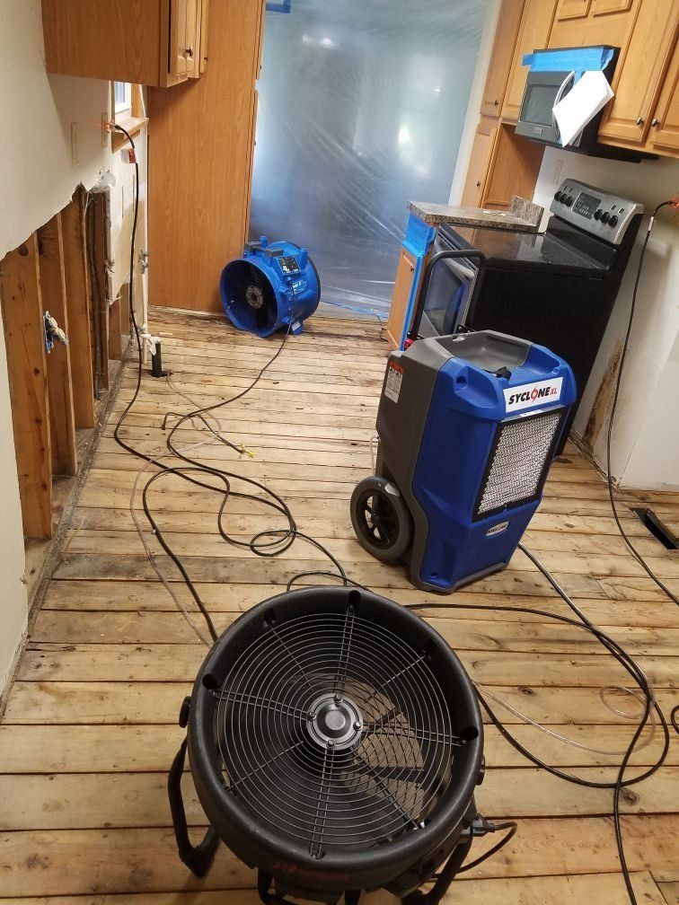 Two industrial fans are sitting on a wooden floor in a kitchen.