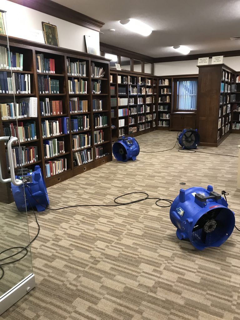 A library with a lot of books on bookshleves and two blue industrial fans on the floor.