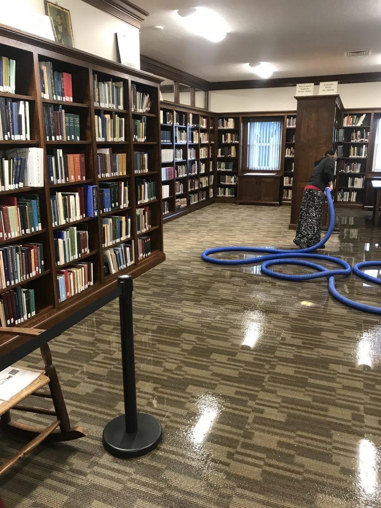 A woman is cleaning the floor of a library with an industrial vacuums cleaner with blue hose.