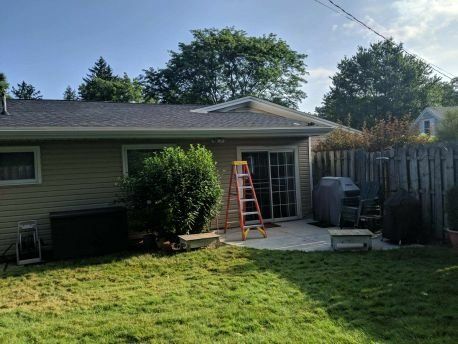 A house with green grass and bush, windows, and patio door with grill.