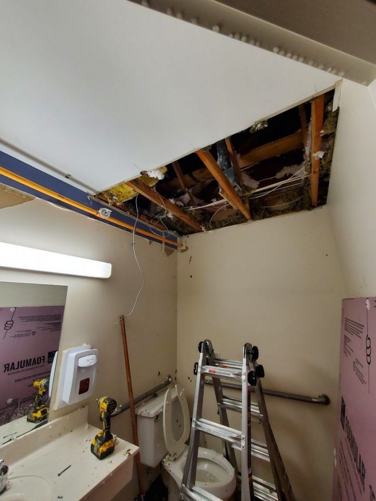 A bathroom with a toilet , sink , mirror, ladder, and damaged ceiling.
