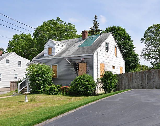 A white house with a green roof and boarded up windows from damage. 