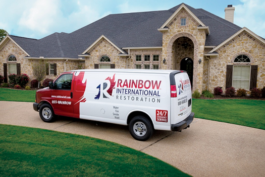 A Rainbow International Restoration van is parked in front of a large brick house.
