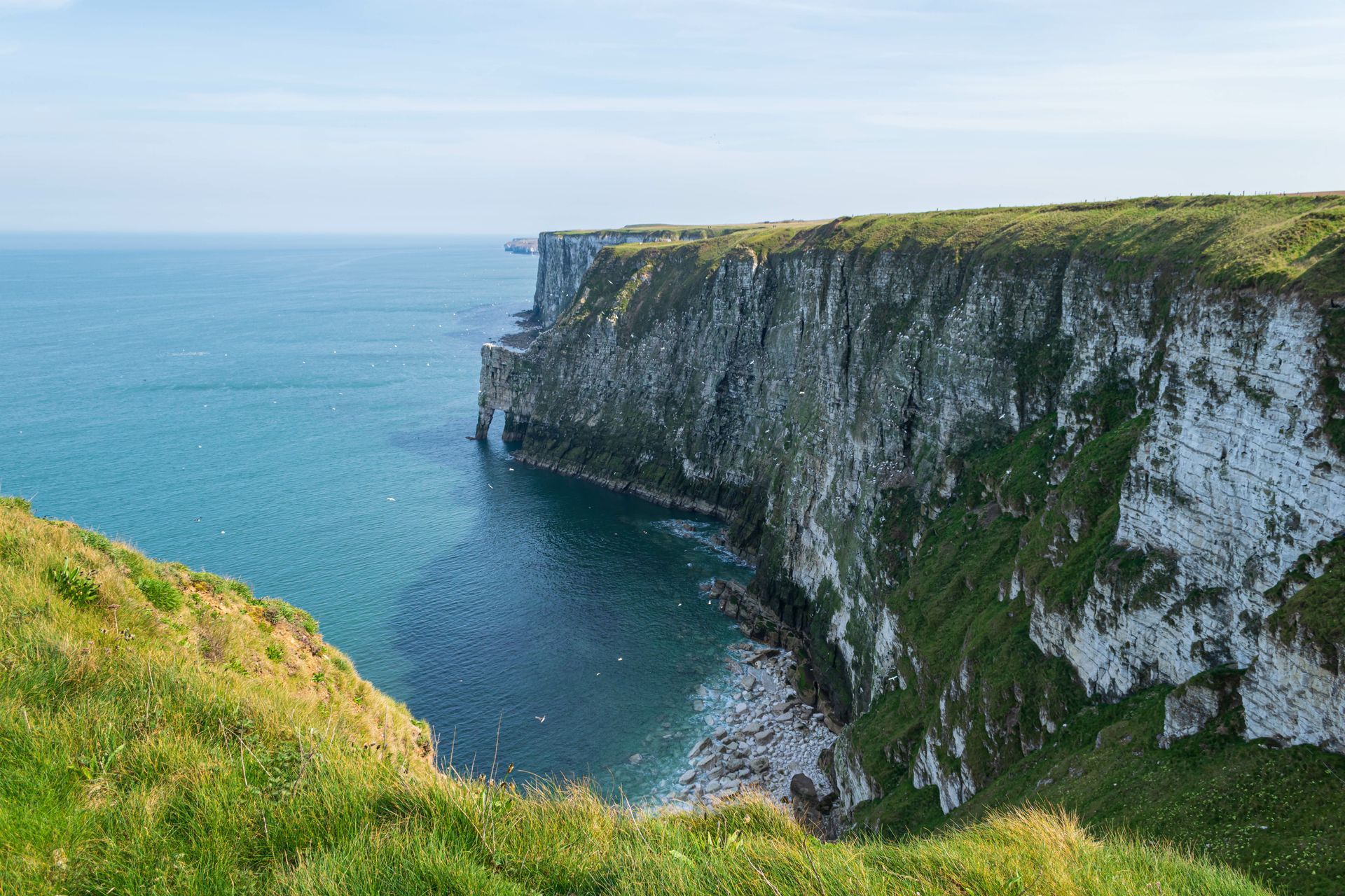 Cliffs overlook a turquoise sea under a blue sky. White chalk cliffs are topped with green grass.