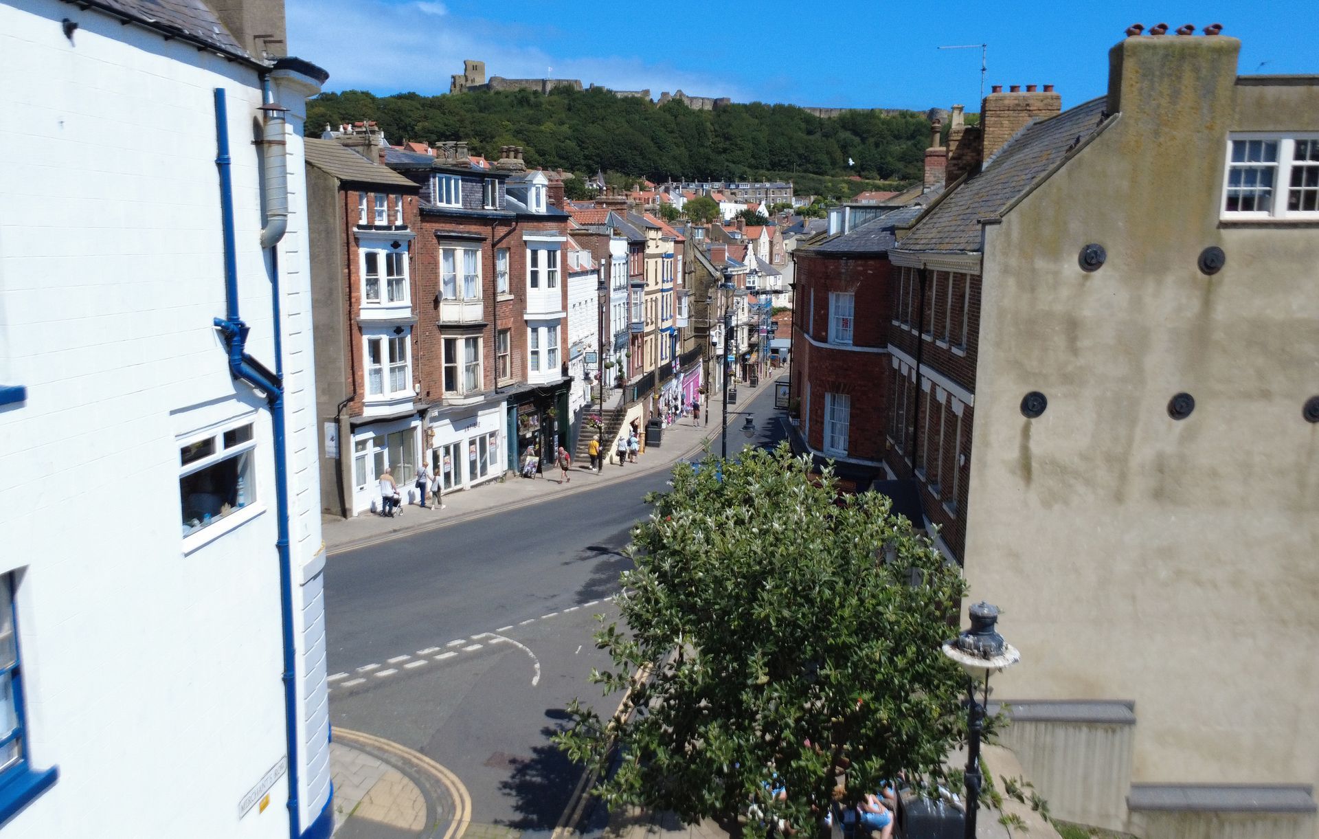 Street view in Scarborough, UK, with buildings lining a road. A castle sits atop a green hill in the background.