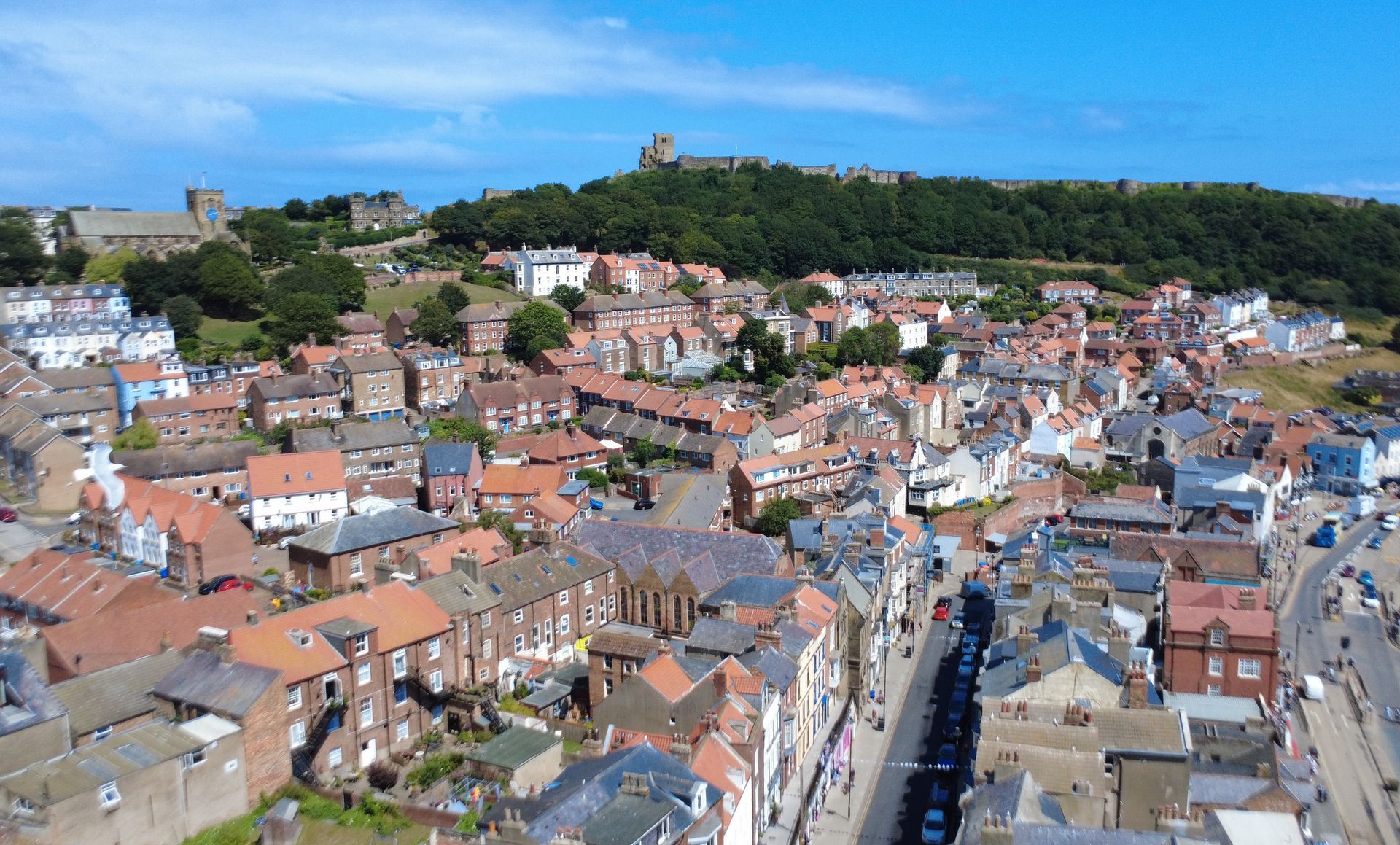 Aerial view of a town with red-roofed buildings, streets, and a castle on a hill under a blue sky.
