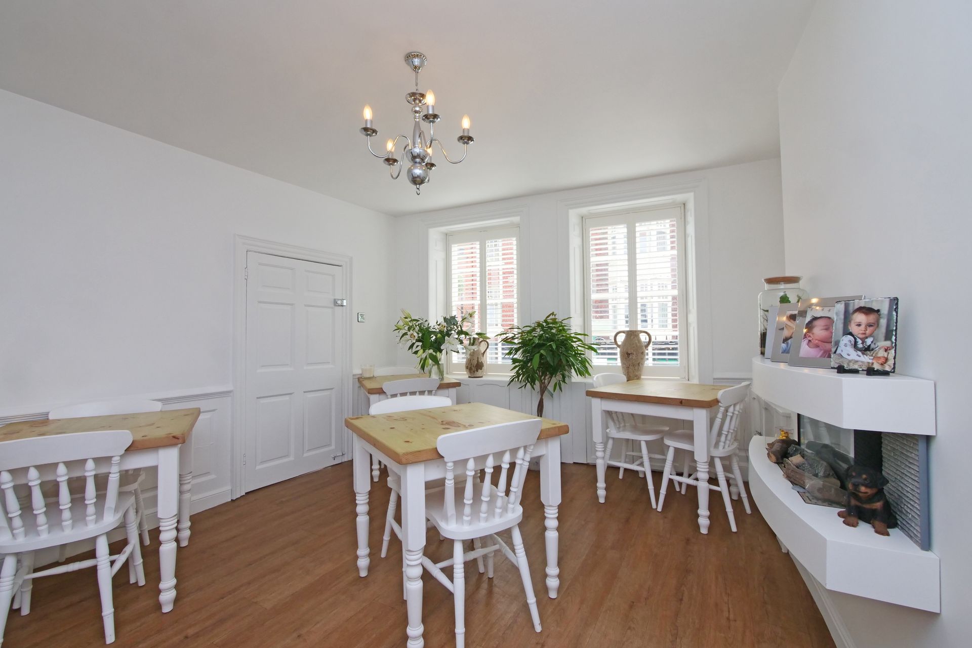 A bright dining area with white walls, wooden tables and chairs, a chandelier, and a fireplace.