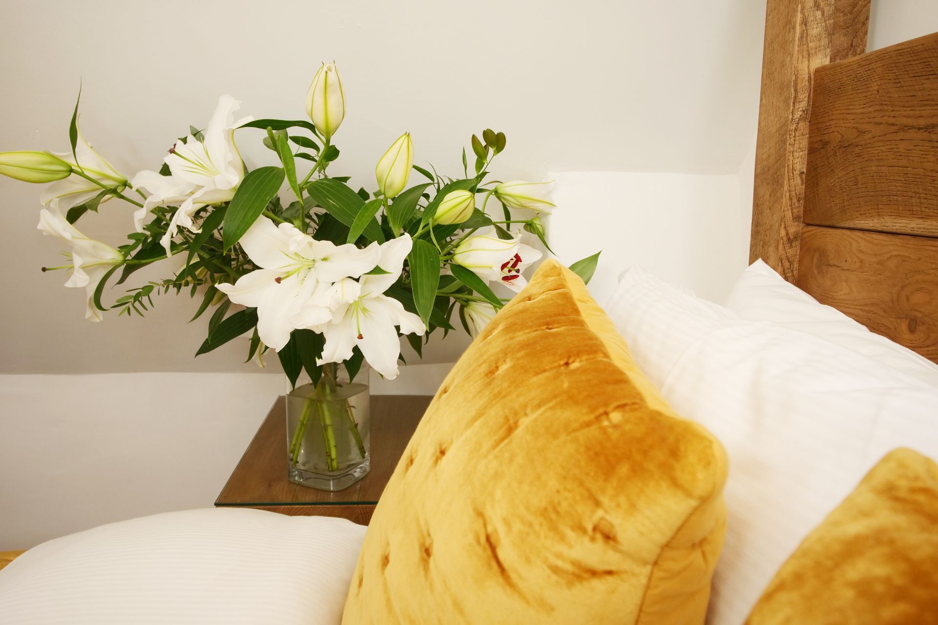White lilies in a vase on a nightstand next to a bed with yellow and white pillows. Wooden beam in the corner.