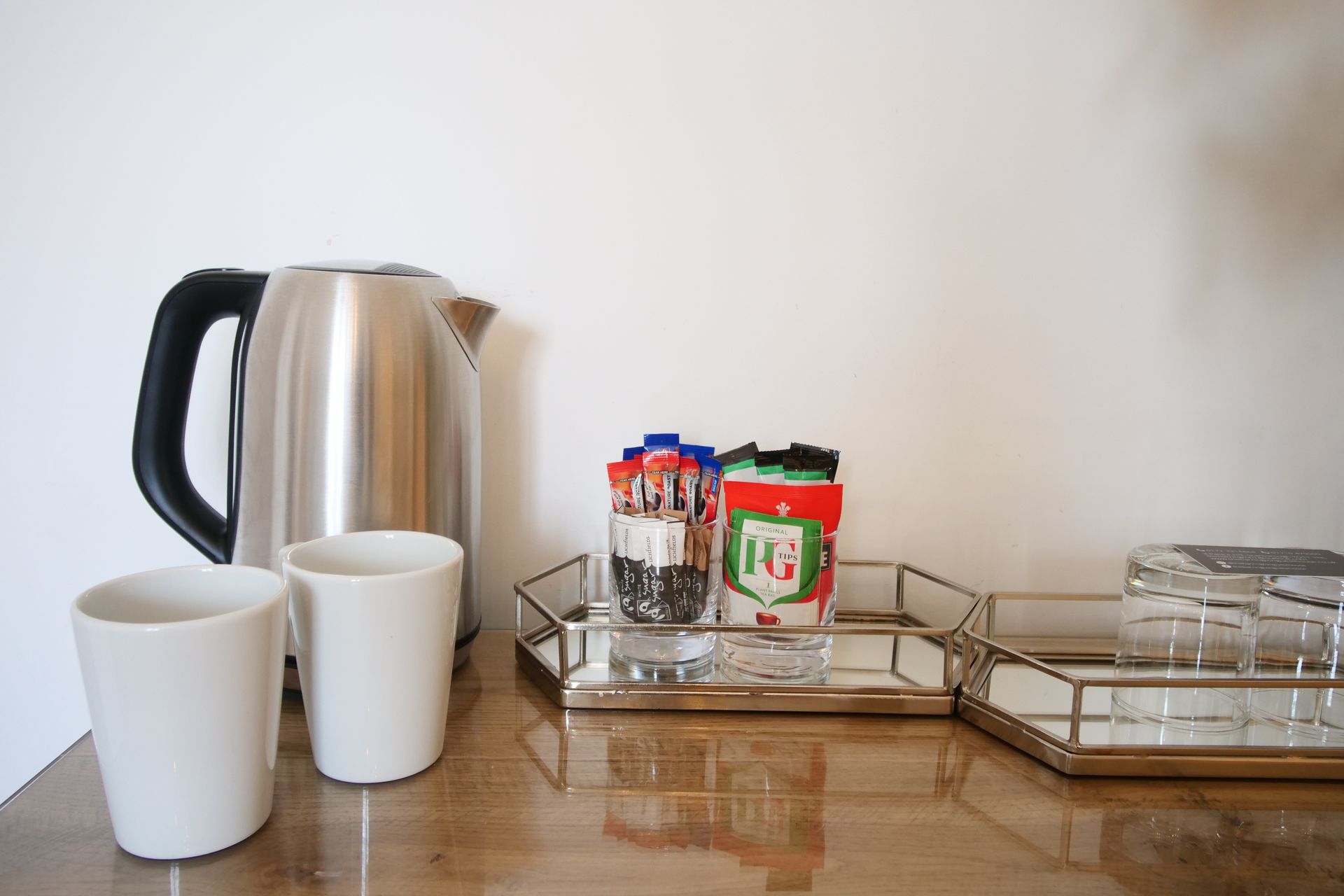 An electric kettle, two white mugs, and coffee supplies are on a wooden surface in front of a white wall.