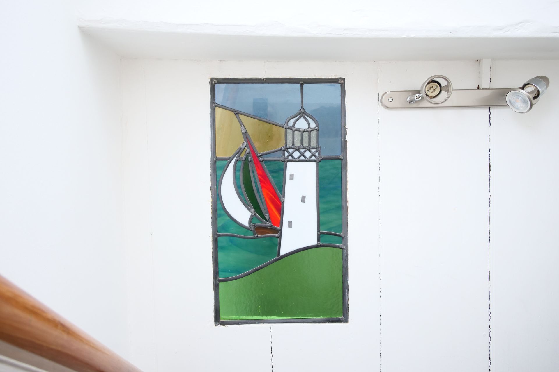 Stained glass window with a white lighthouse, sailboat with red sail, and green sea and land. Set in a white doorway.