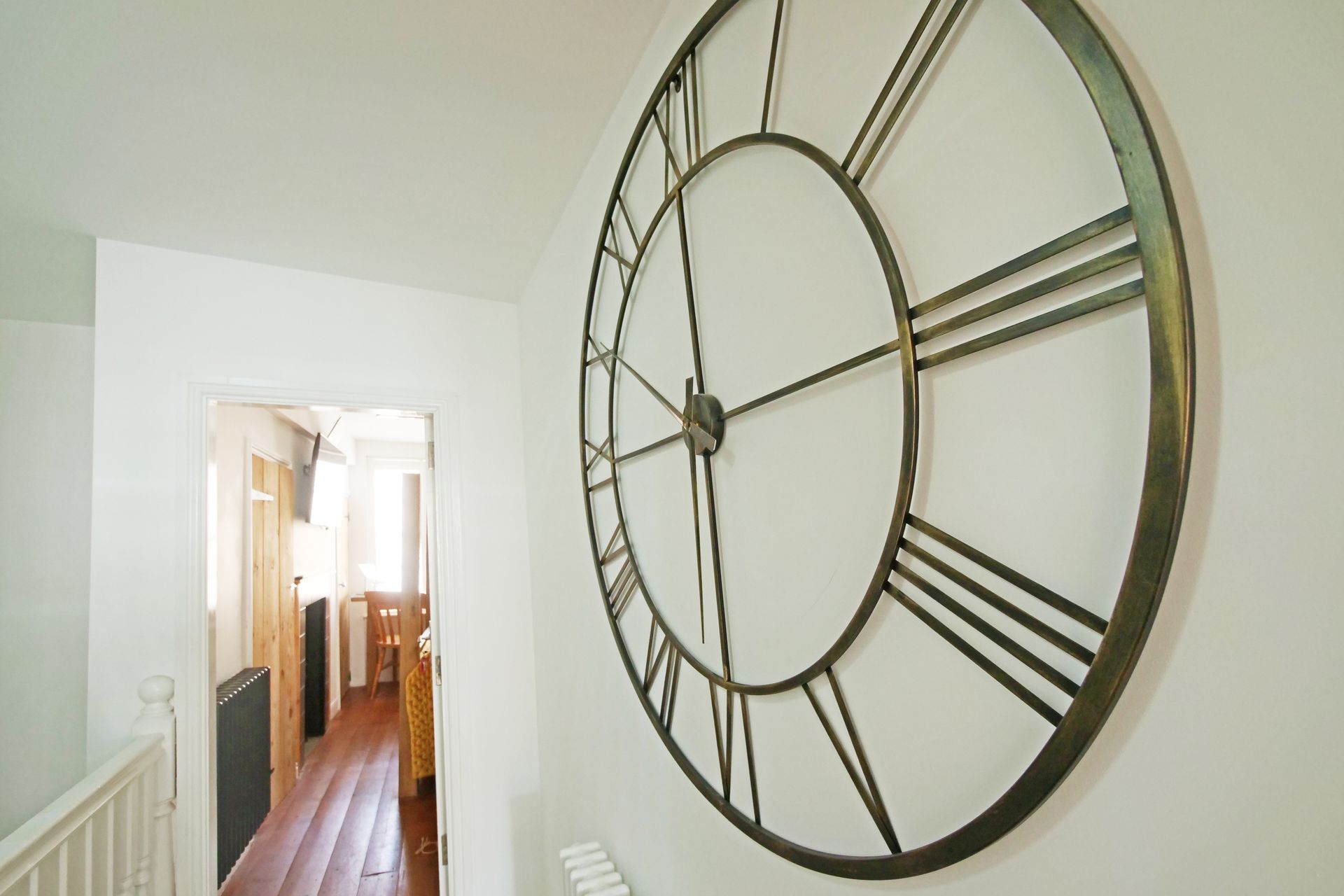 Large clock with Roman numerals on a white wall in a hallway. A doorway leads to a room with a radiator.