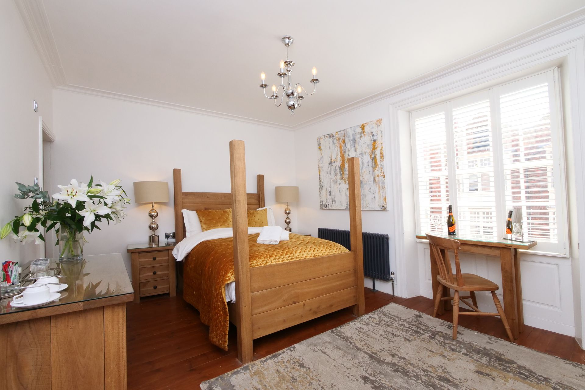 A cozy bedroom with a four-poster bed, wooden furniture, a desk by a window, and a vase of flowers.