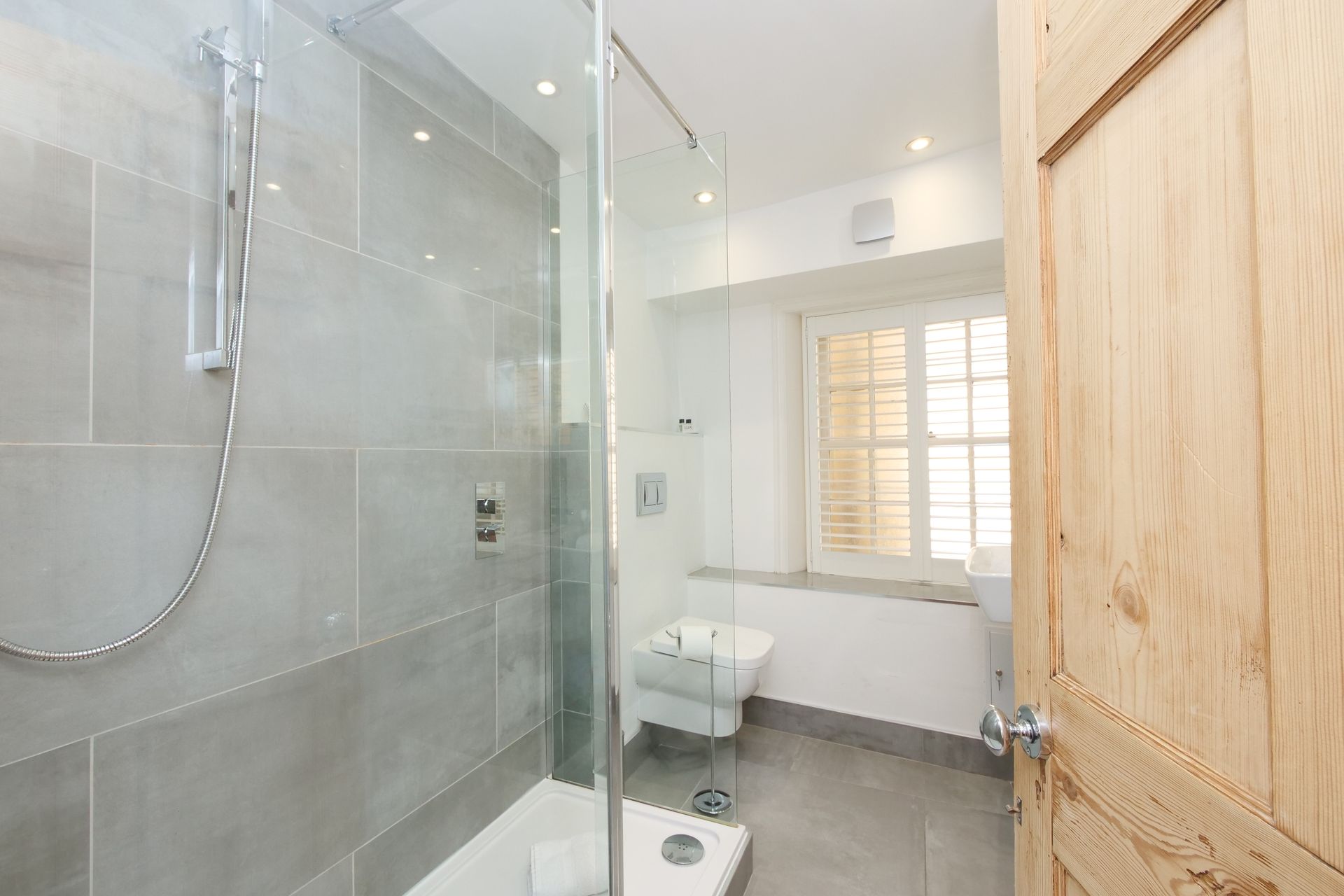 A modern bathroom with a glass shower, gray tiles, a window with shutters, and a light wooden door.