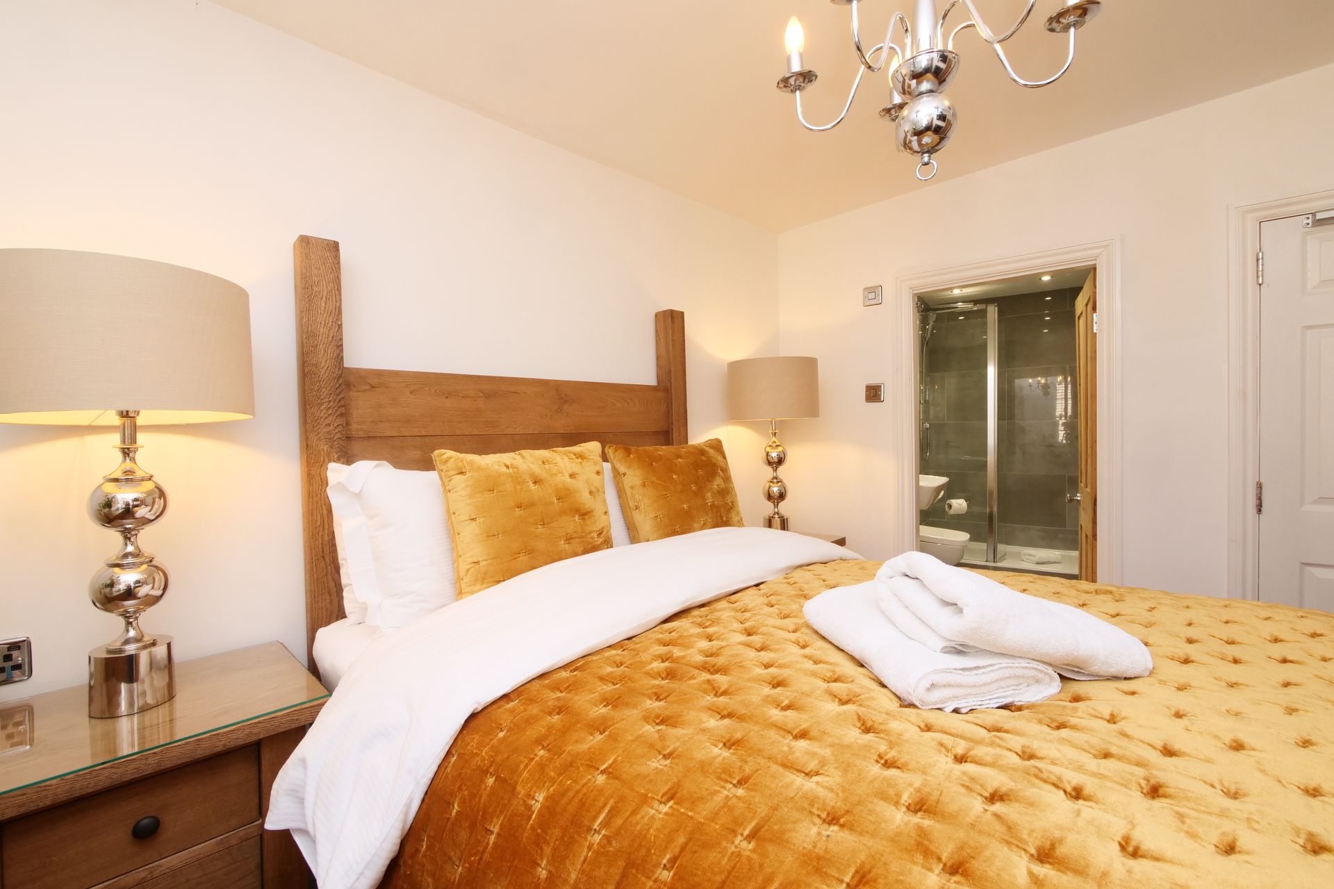 Bedroom with a mustard-coloured bedspread, matching pillows, wooden headboard, and a chrome chandelier. A bathroom is visible
