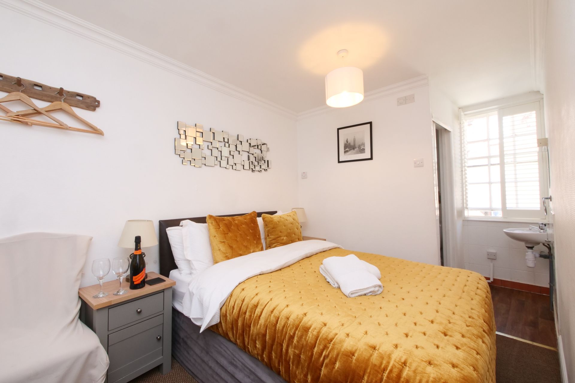 Cozy bedroom with a gold comforter, bedside table, chair, and small sink. White walls and a window provide natural light.