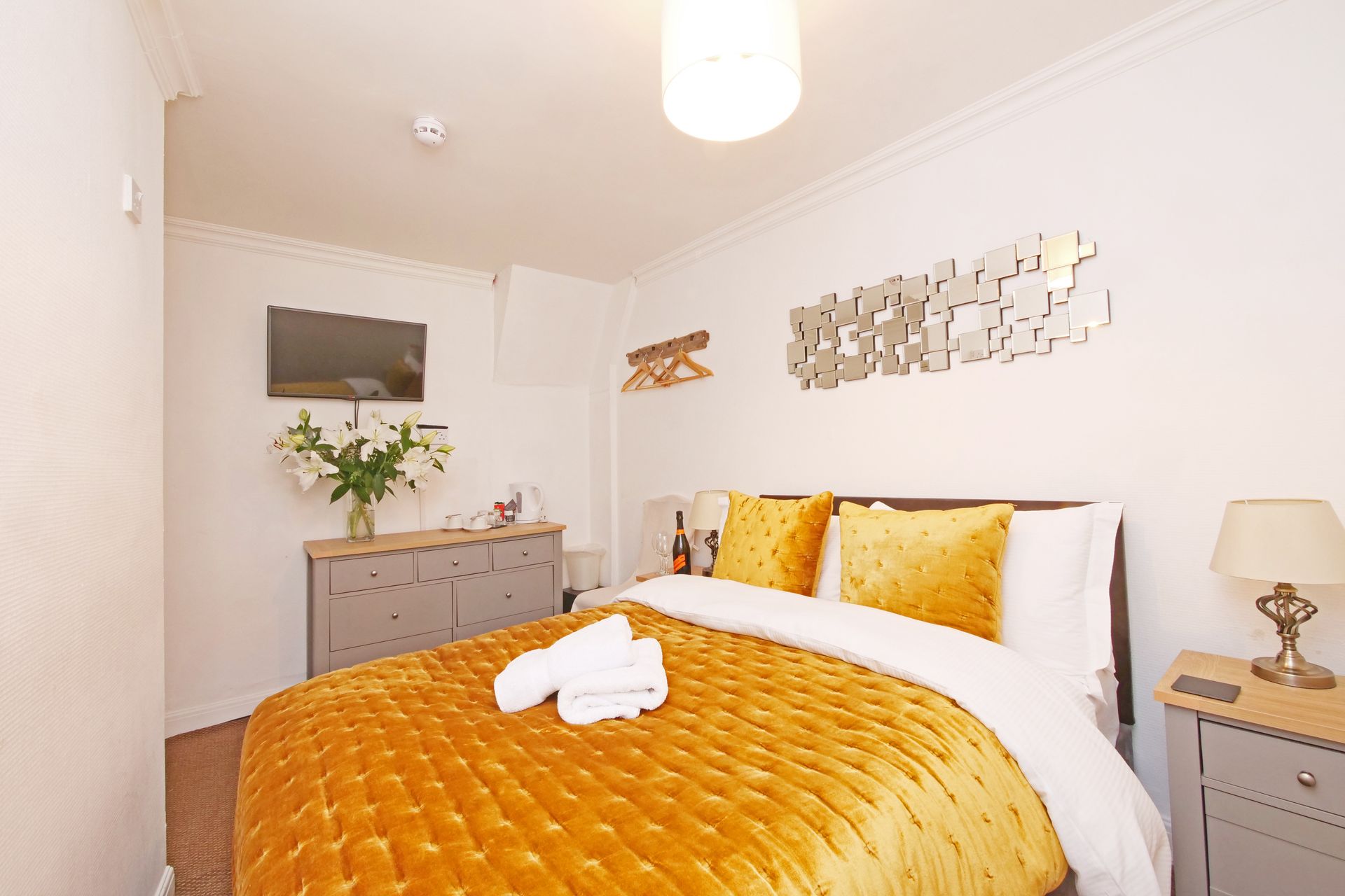 A cozy bedroom with a gold bedspread, bedside table, and a wall-mounted TV. There's a dresser with flowers