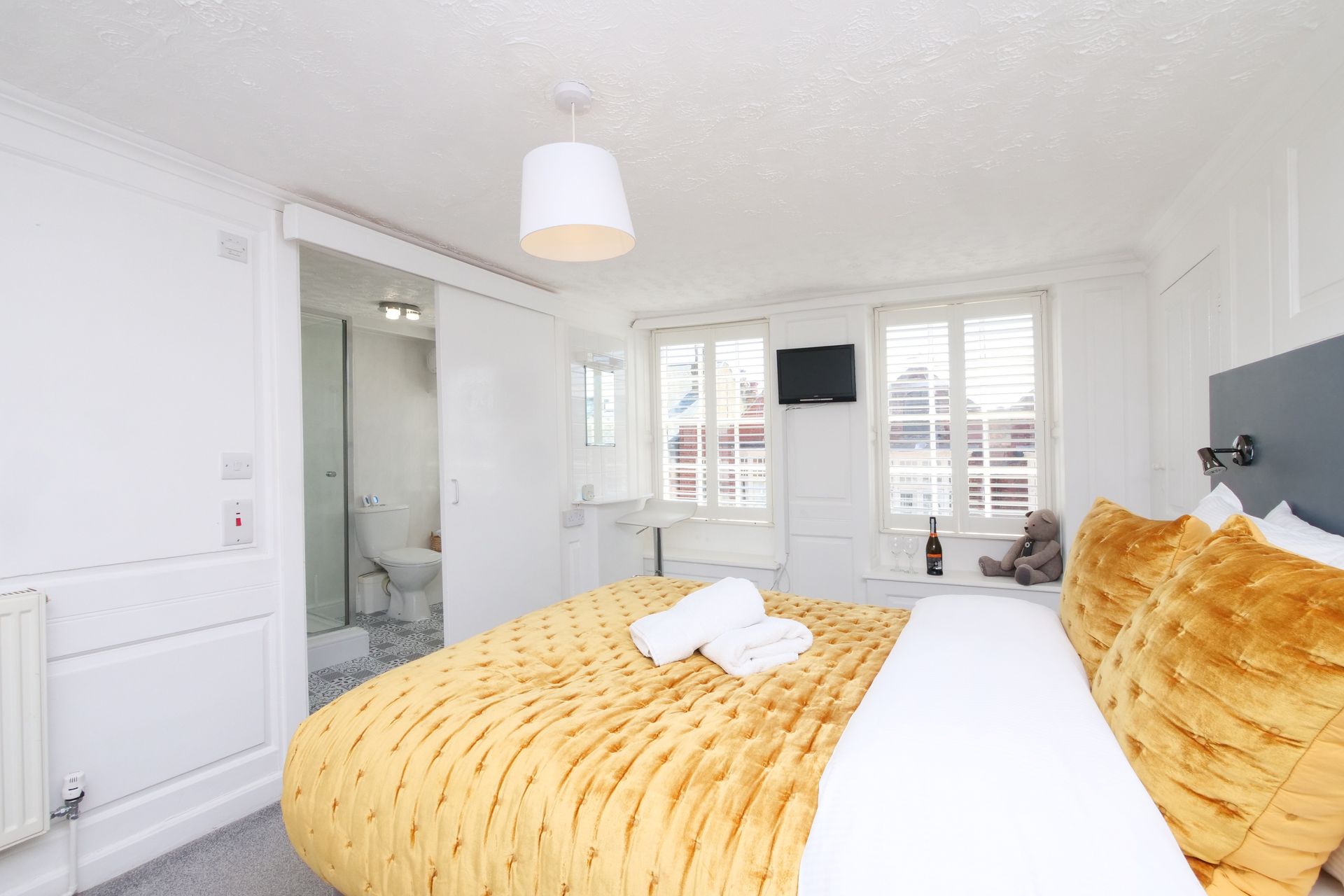 A bright bedroom with a yellow bedspread, two windows, a small TV, and a partially visible bathroom.