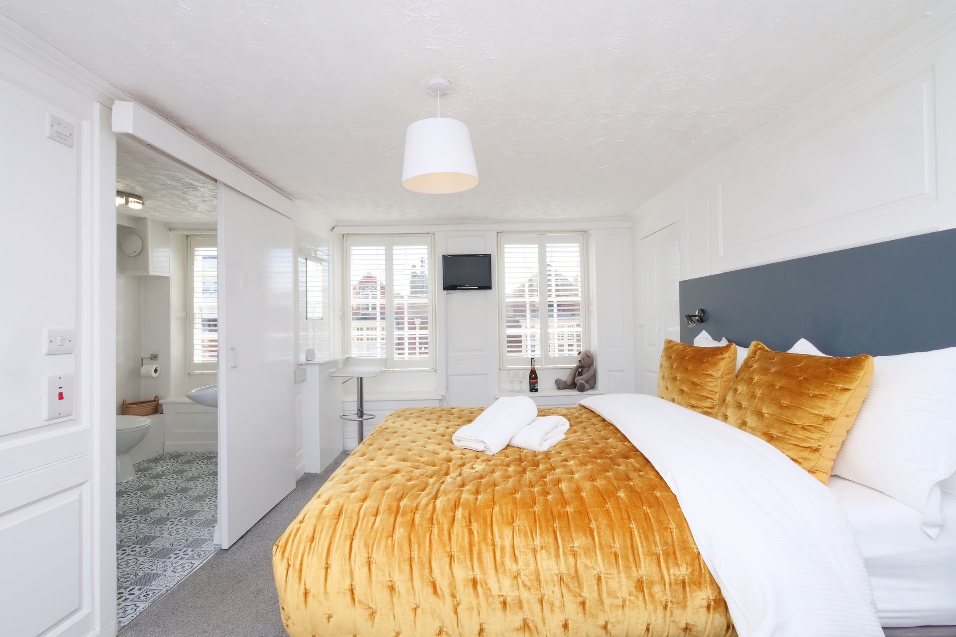 A hotel bedroom with a gold bedspread, two windows, and a bathroom visible through a sliding door.