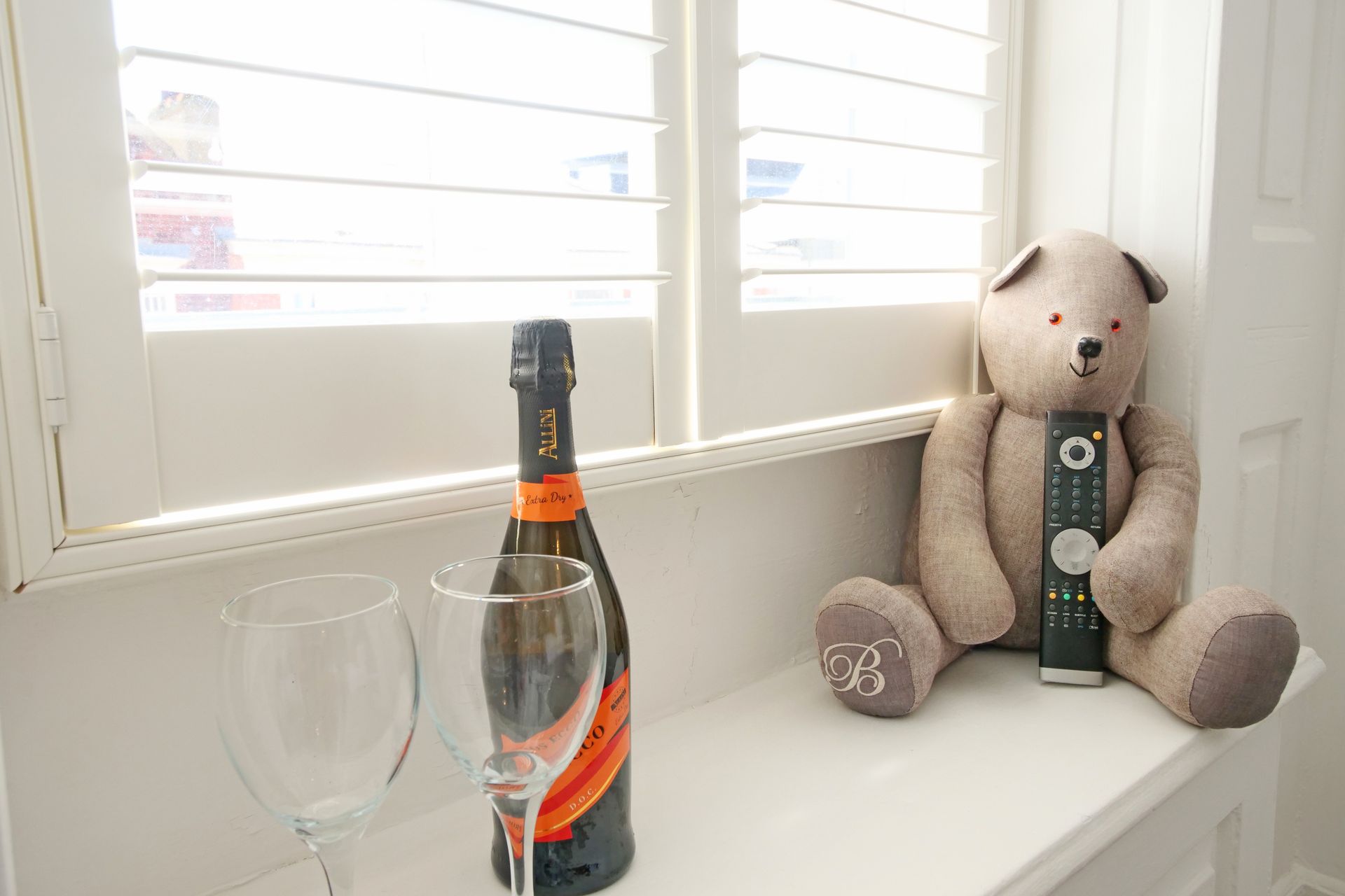 A teddy bear sits on a windowsill, holding a remote, next to a champagne bottle and two empty glasses.