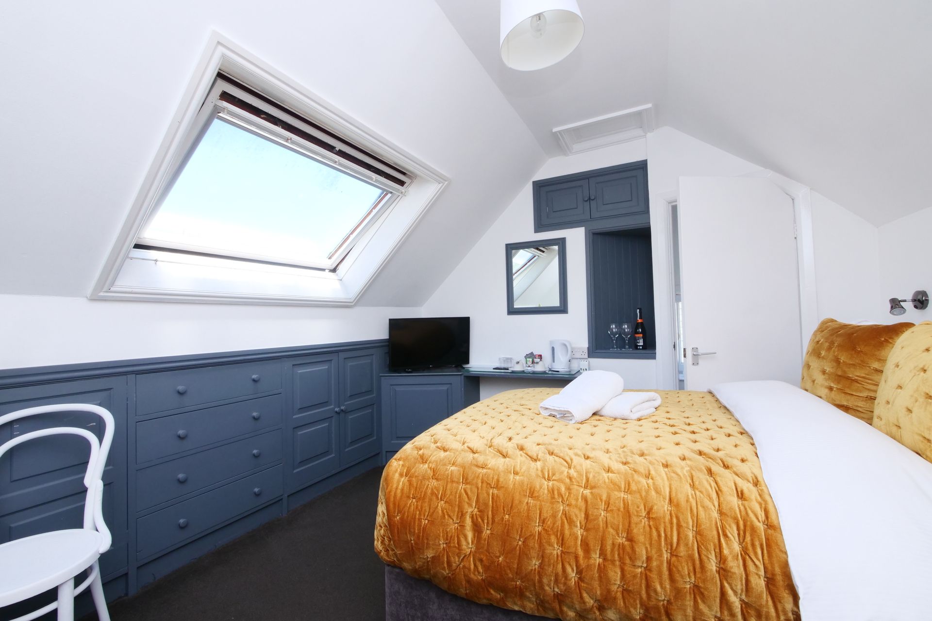 Bedroom with angled ceiling, skylight, mustard-colored bedspread, built-in gray cabinets, and white chair.