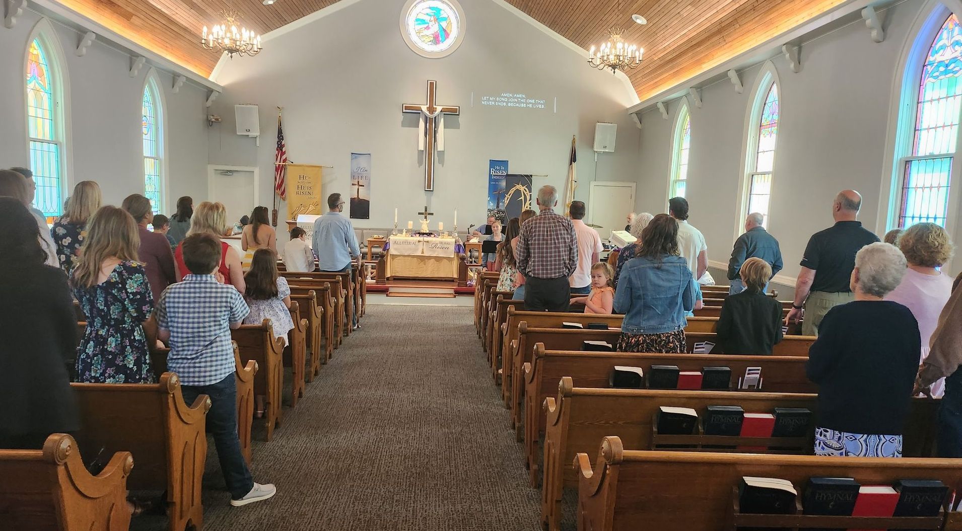 People standing in a church during a service, facing altar, with pews and stained glass windows.