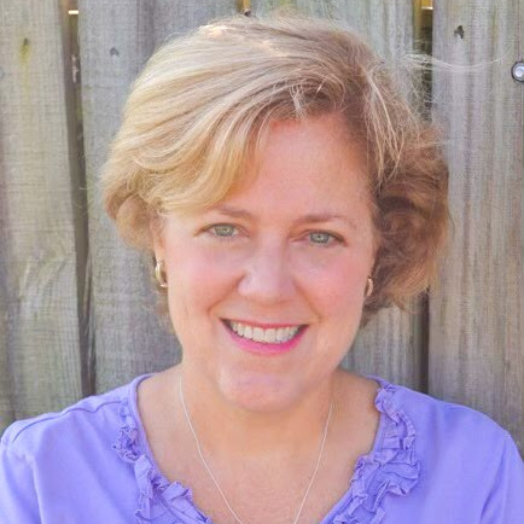 Woman with blonde hair, smiling, wearing a purple shirt, in front of a wooden fence.