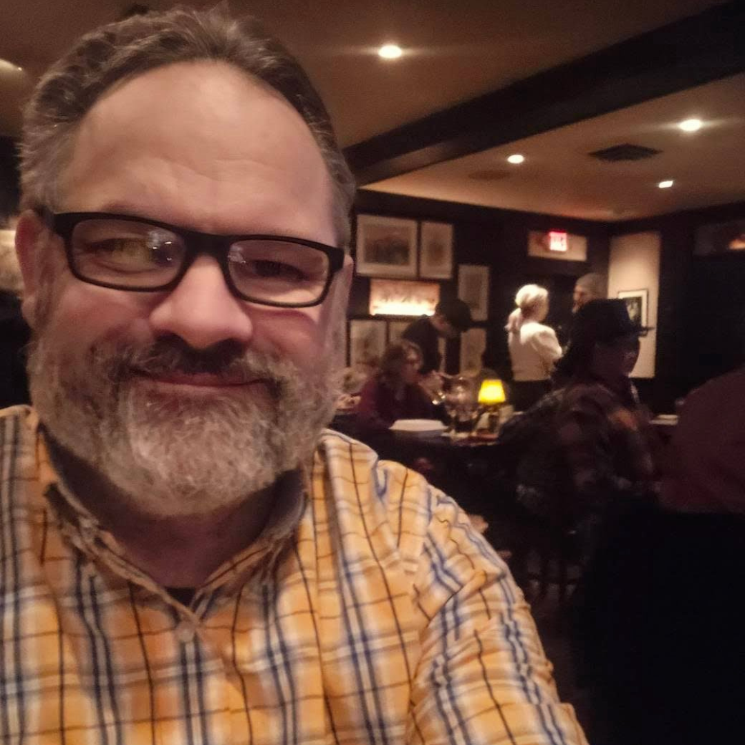 Man with glasses and beard in a plaid shirt smiling at a dimly lit bar with other patrons.