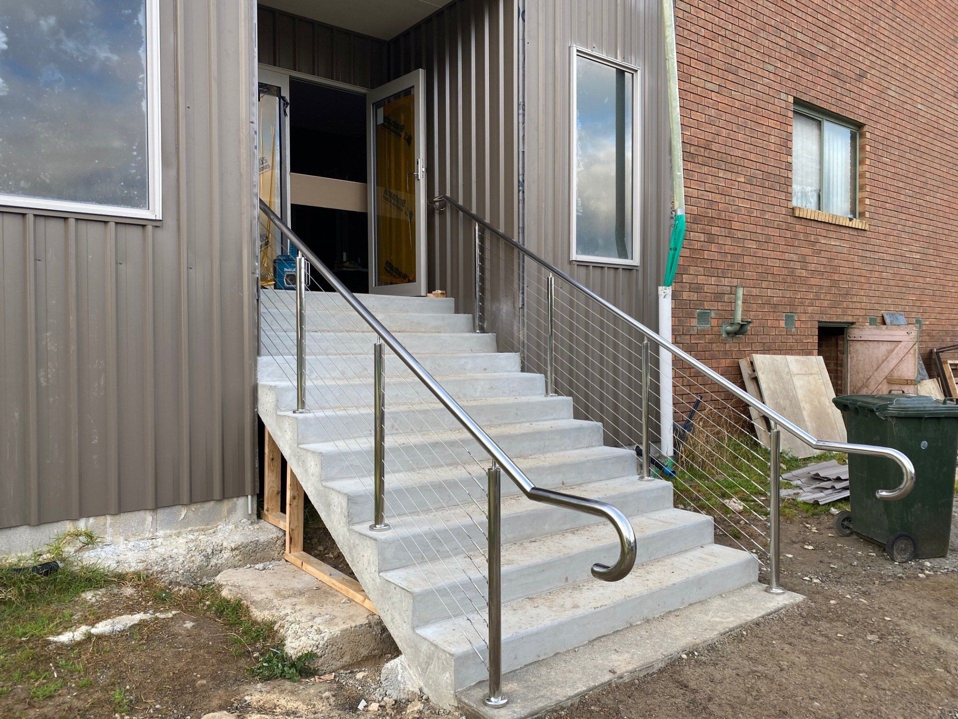 Gray metal stairs with stainless steel handrails lead up to a door on a corrugated metal building, next to a brick wall.