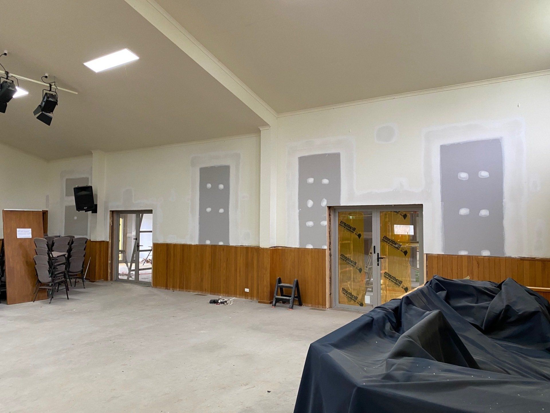 An empty room under renovation, with wood paneling, drywall, and a covered object in the foreground.
