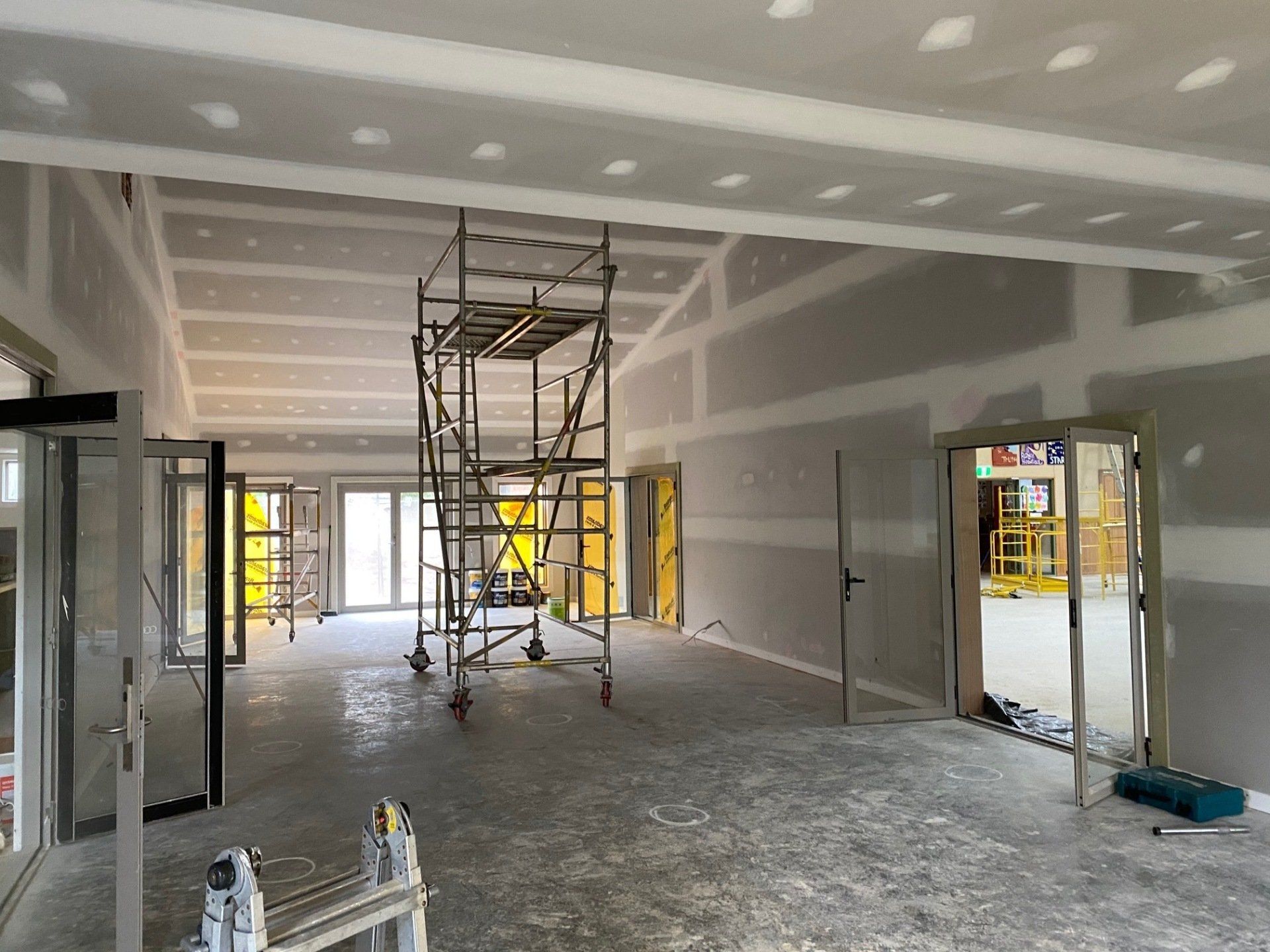 Interior of a room under construction, with drywall walls and ceiling, scaffolding, and glass doors.