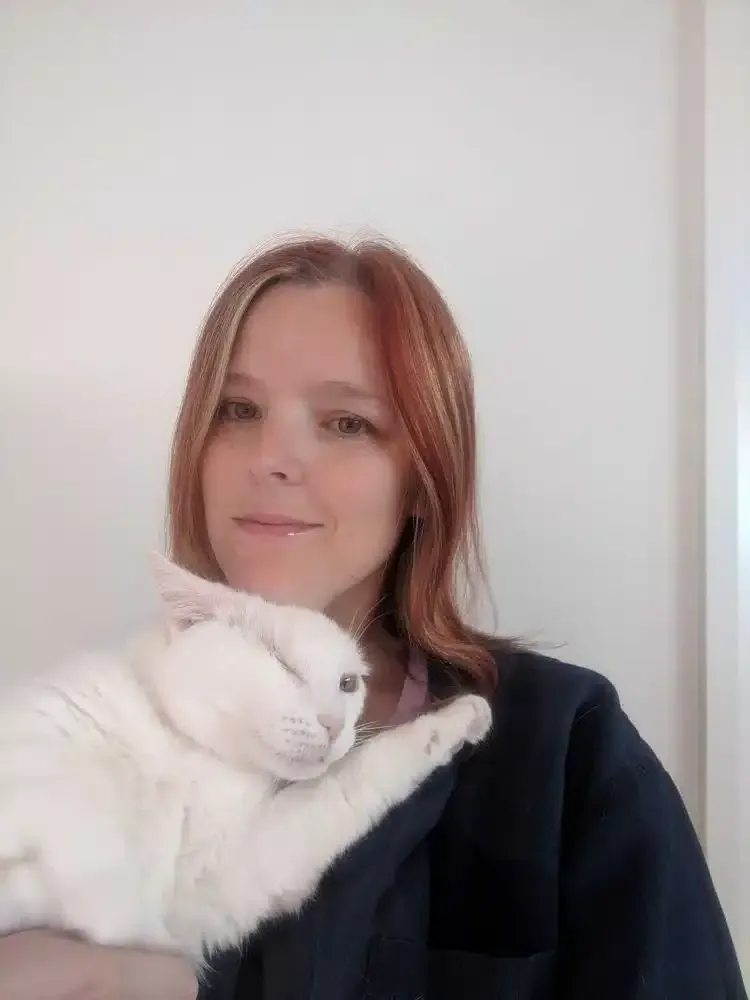 Woman holding a white cat. Both looking off-camera. The woman has red-toned hair and is smiling slightly. White wall.