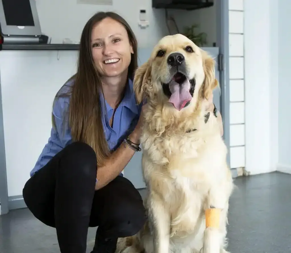 Woman with long brown hair kneels next to a golden retriever with a bandage on its leg; they smile.