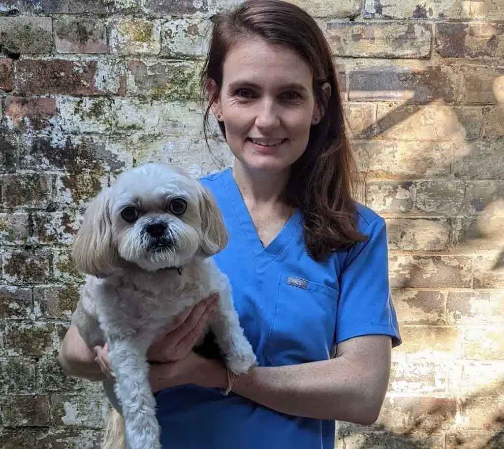 Woman in blue scrubs holds a small white dog, smiling in front of a brick wall.