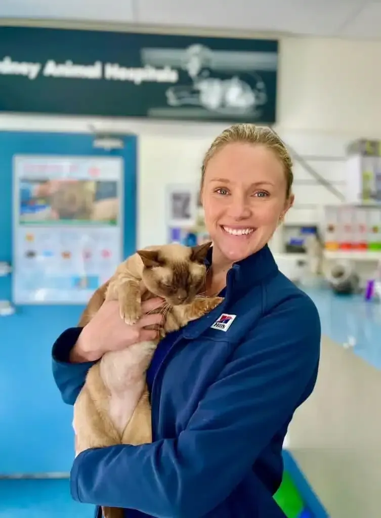 Woman in blue jacket smiles, holding a beige and brown cat in a veterinary clinic.