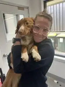 Woman in glasses holding a brown and white dog; smiling indoors near a window and bins.