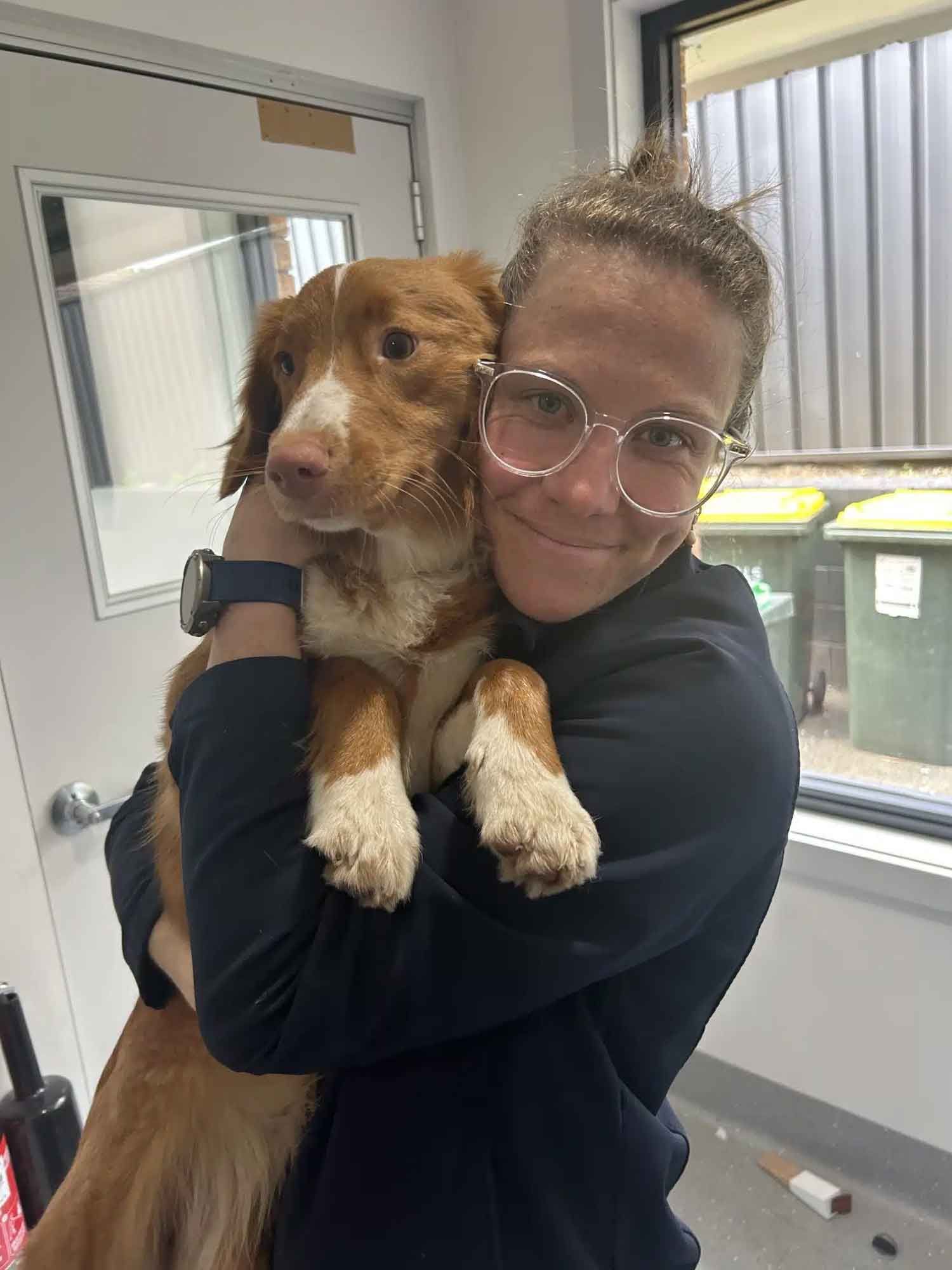 Woman hugging a brown and white dog inside a building. Both are smiling.