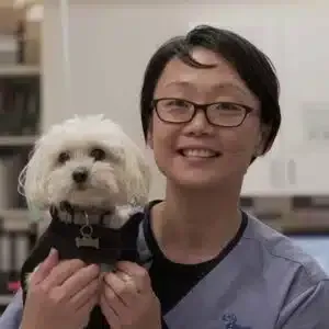 A woman in glasses holds a small, white dog, both smiling. They are indoors, likely a clinic.