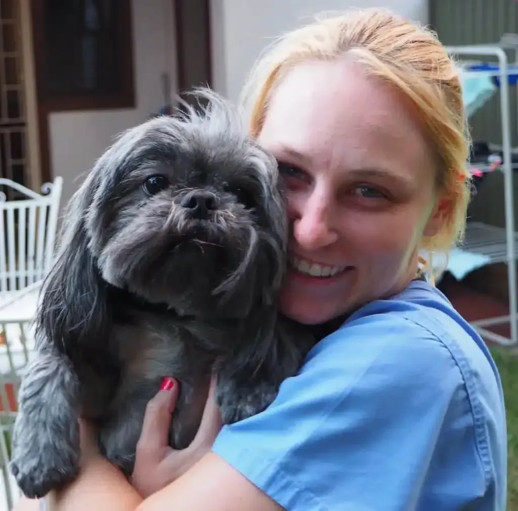 Woman in blue shirt holding a gray Shih Tzu dog; both smiling outdoors.