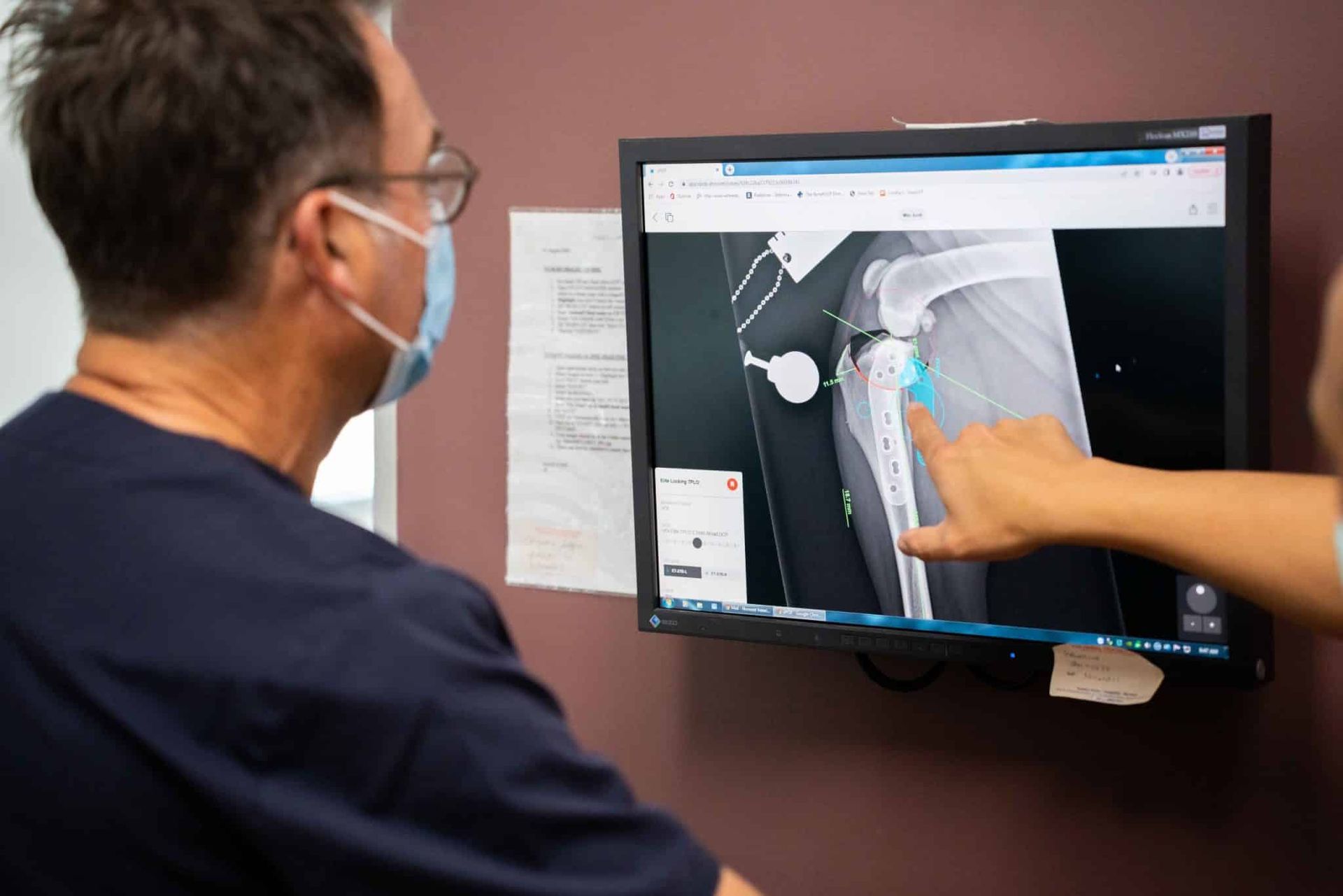 Doctor in mask looks at computer screen with x-ray, another person points at the image on screen.