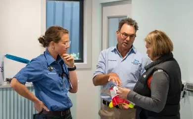 Three people, two in scrubs, talking in a brightly lit room. A woman holds a red toy.