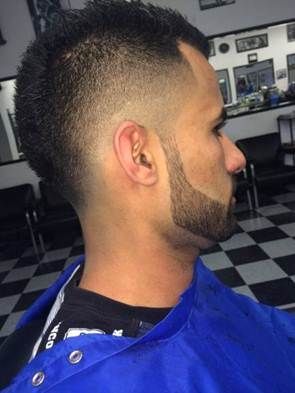 Man with dark hair and a faded mohawk-style haircut, also a short beard. He is wearing a blue barber's smock.