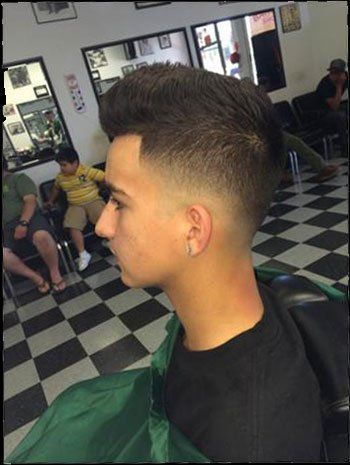 Man with a fade haircut in a barbershop, posing. Black and white checkered floor, other people visible.