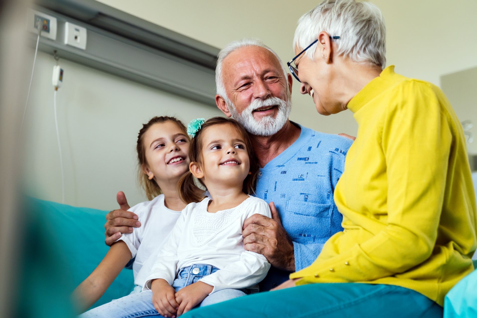 A smiling grandfather in a hospital gown hugs two young girls while a woman in a yellow sweater looks on.