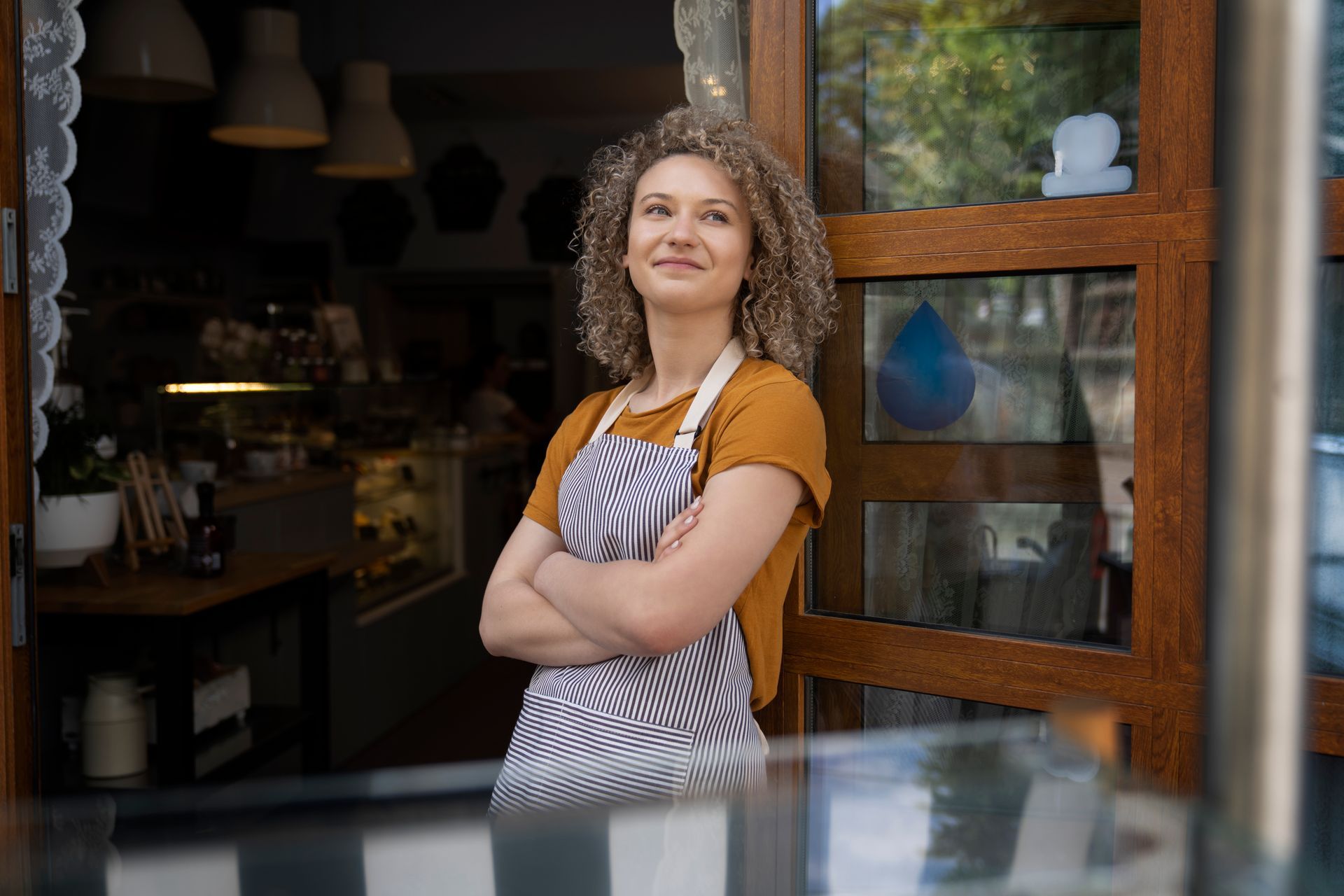 A person with curly hair wearing an apron and mustard top stands with arms crossed in the doorway of a cafe.
