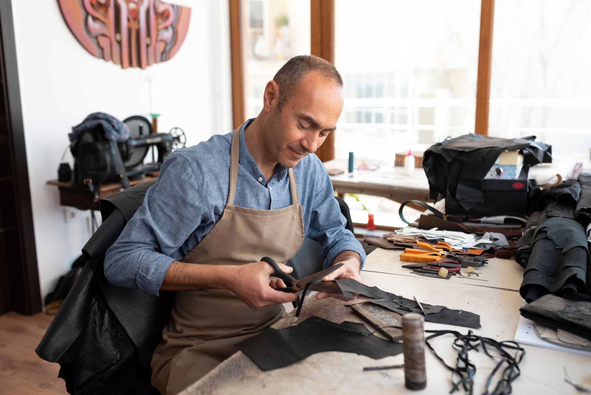 A person wearing a tan apron cuts a piece of black leather with scissors in a bright, organized workshop.