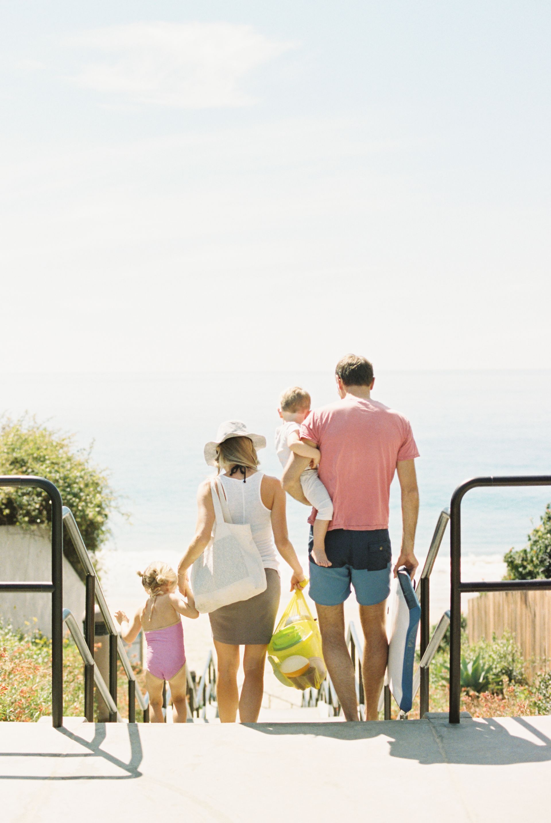 A family of four walking down stairs toward a sunny beach while carrying bags and towels.