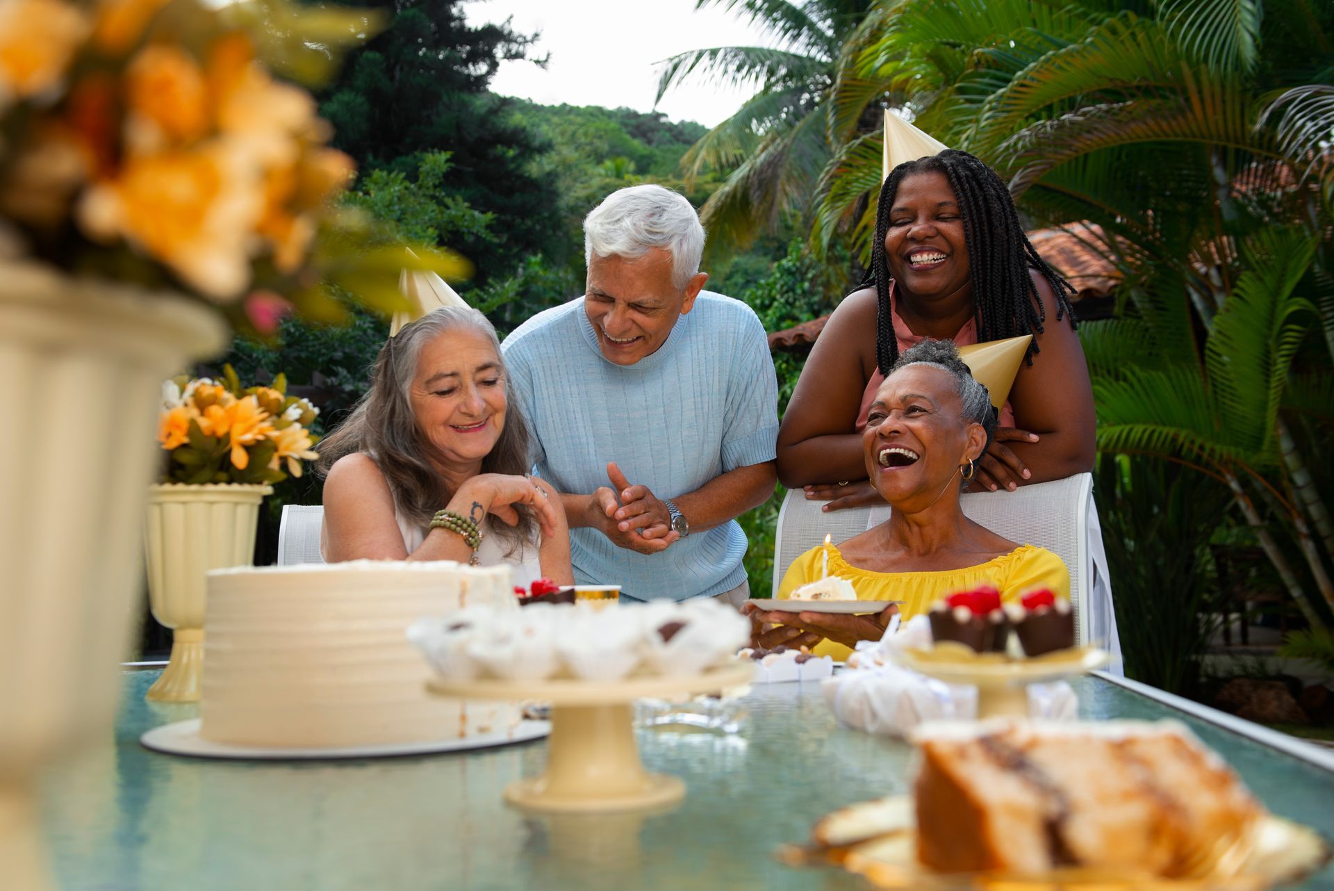 Four people laughing at an outdoor table with a cake and treats.