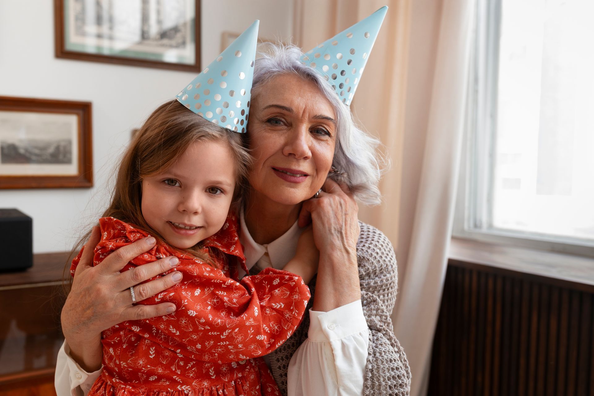 A child and an adult wearing light blue party hats embrace while smiling in a softly lit room with framed wall art.