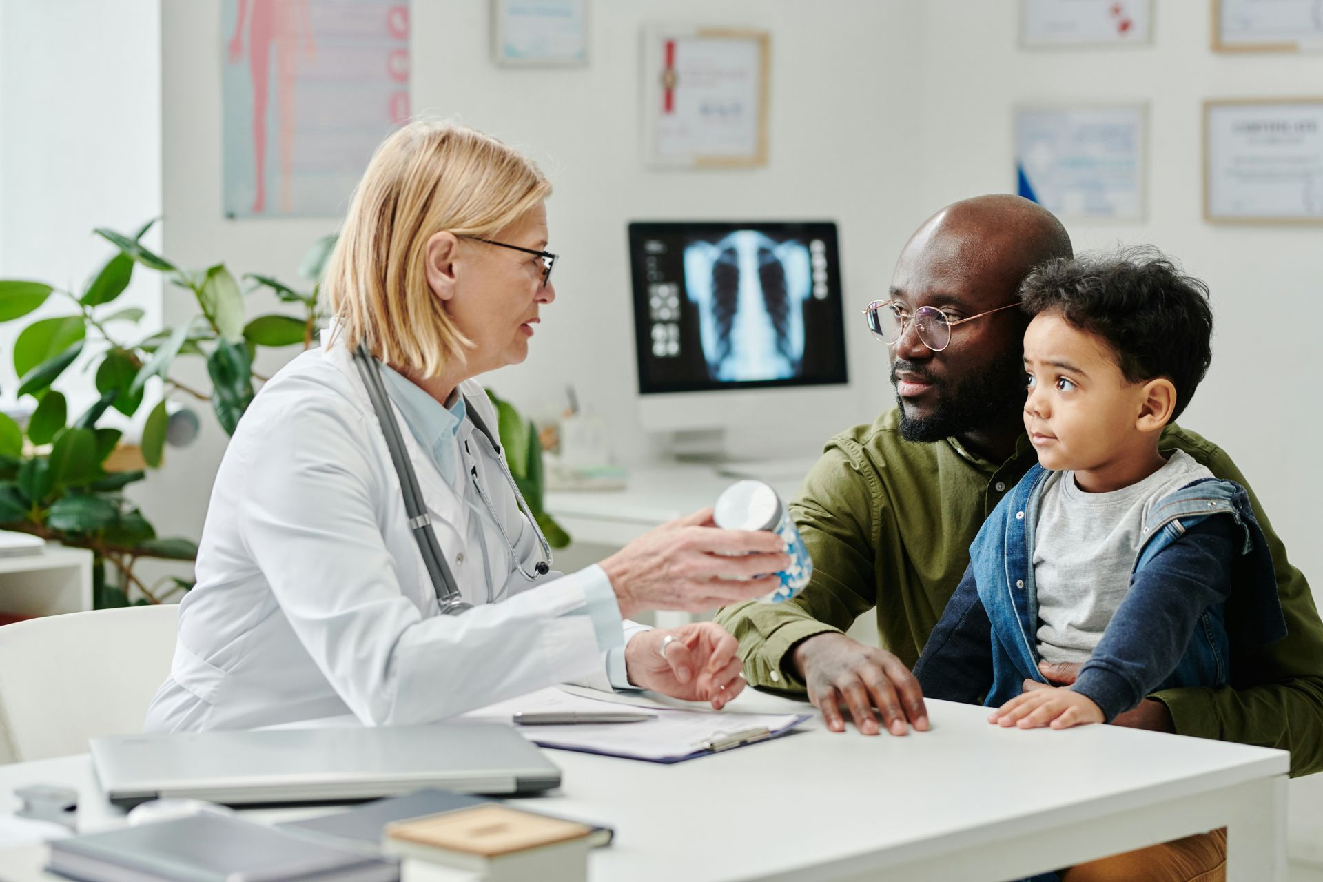 A doctor sits at a desk in an office, discussing medication with a man and a young child.