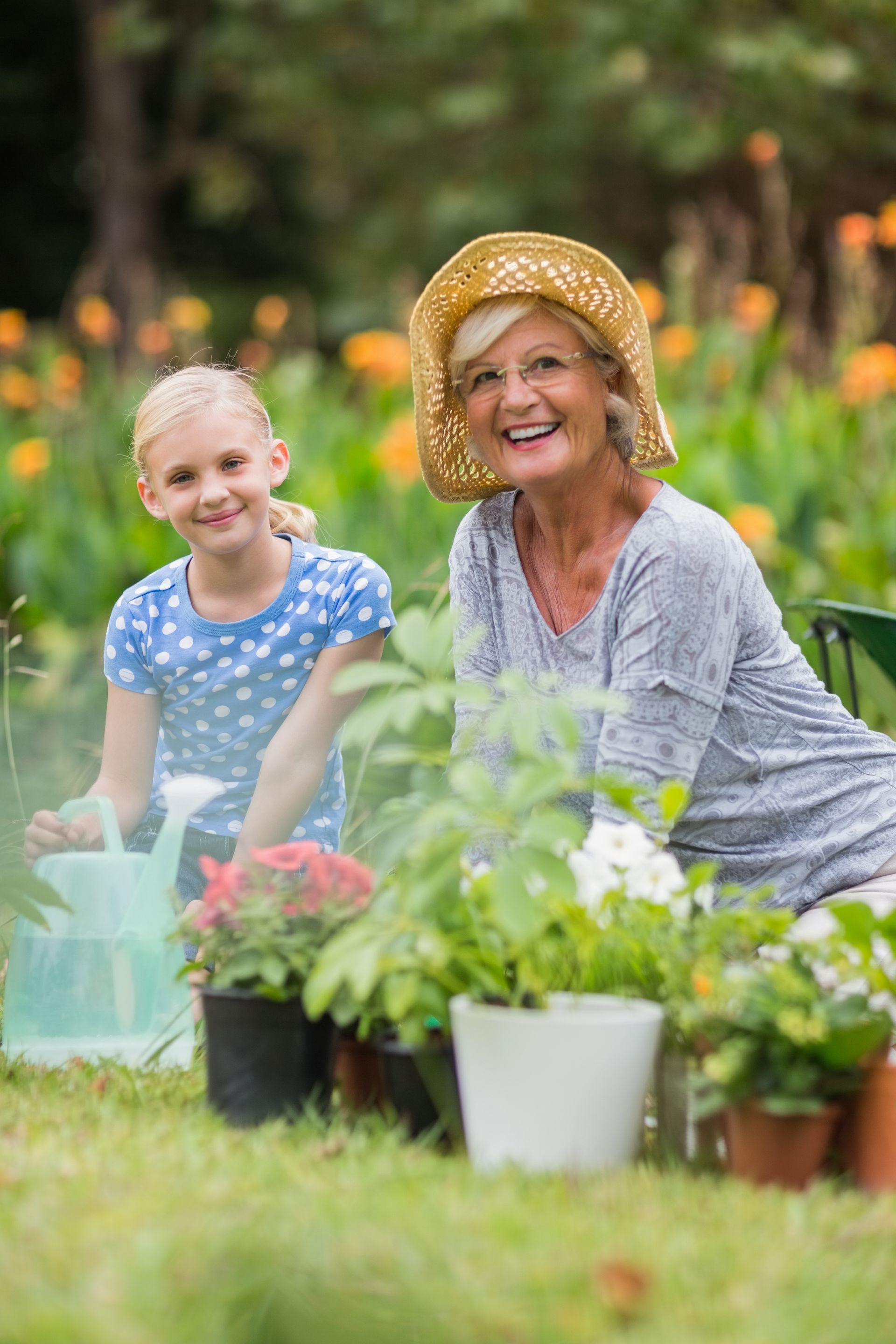 A person in a sun hat and a child gardening together with potted plants and a watering can in a grassy area.